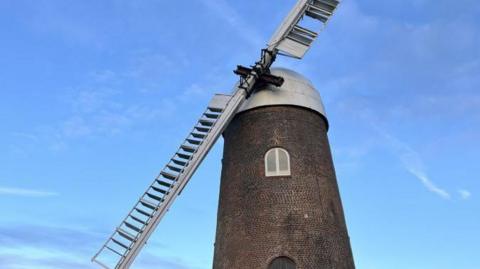 A brick windmill with a small white window, a white roof and two white wooden sails against the background of a blue sky on a sunny day. 