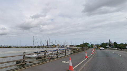 Google view of the A180 flyover at Alexandra Dock in Grimsby. There are cones laid out along one side of the road next to a cycle lane. There are a number of boats visible in the distance.