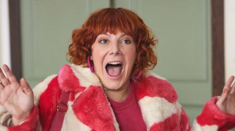 Sophie Willan starring in series two of Alma's Not Normal - she has red short curly hair and a pink and white striped fake fur coat