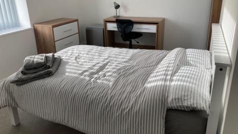 A photo showing a white bedroom with a grey and white striped bed, with towels folded at the foot. There is a brown and white desk against the far wall, with a black office chair and a chest of drawers to the left.