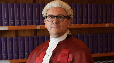 Lord Colbeck looking at the camera. He is wearing a white wig and a long red robe with red crosses on it. He has a white collar on. He is standing in front of a bookshelf containing books with blue spines.
