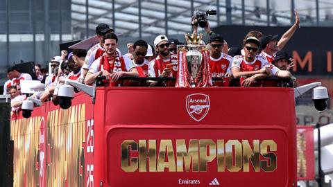 Arsenal men's team on top of a red double-decker bus saluting the crowd as they parade with their Premier League trophy