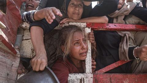 A young girl looks distressed as she attempts to get food, surrounded by others. One person holds a bowl.