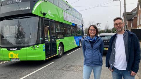 Two people are standing the right of the picture. A man and a woman. To their left is a green bus.