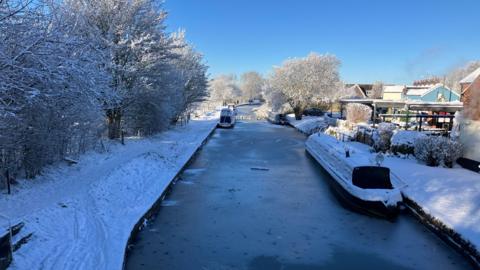 A frozen canal with two snow-covered narrow boats moored up either side. The houses to the right are covered in snow as are the towpath and trees on the left.