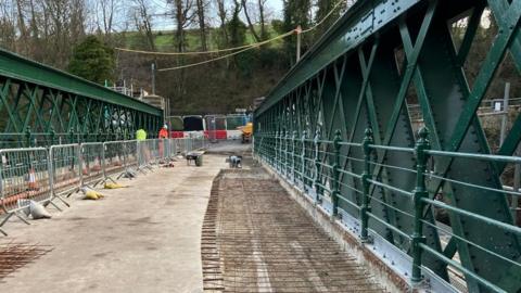 Wolsingham Causeway Bridge is narrow and has tall green steelwork on either side. There are temporary barriers denying access to it and part of its surface is exposed with metal bars underneath. There are two workers in high-visibility clothing at the end of the bridge. 