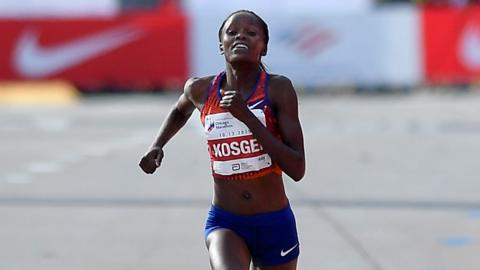 Brigid Kosgei, wearing a red vest with her name on it and blue shorts, swings have arms and grimaces as she struggles towards the finish line in the Chicago Marathon