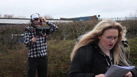 A young girl with long blonde hair, she wears reading glasses while holding a script in a field. There is a man holding a camera behind her wearing a white hat and black and white checked shirt.