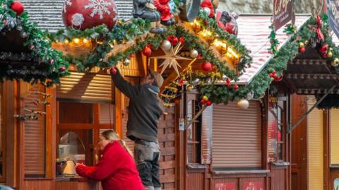 A man and a woman work on their stall at a Christmas market in Germany - the woman in a red coat is on the phone while the man tends to his stall