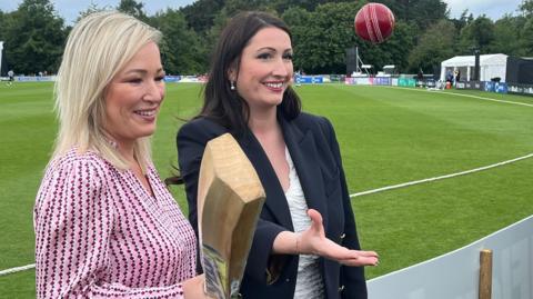 Michelle O'Neill smiling holding a cricket bat and Emma Little-Pengelly beside her throwing up a cricket ball.