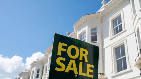 A black sign with 'for sale' in yellow writing printed on it, in front of a white house.