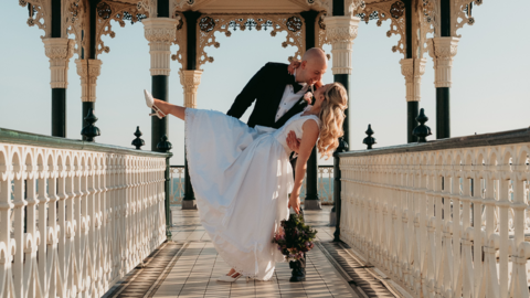 The image shows a groom holding his bride as she dips with one foot in the air and a bouquet in her hand. They are on what looks like a pier and the sky behind them is clear.