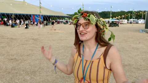 A woman with long dark hair stands on a field with very dry grass. She wears a flower crown on her head.