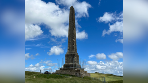 A tall stone monument on a grassy hillside set against a blue sky with some small white clouds