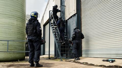 Four people dressed in all black with helmets on are stood around a set of metal stairs next to an industrial building. At the top of the steps someone can be seen using an object on the door with the other people looking at what the person is doing.