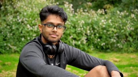 Mridul is sat in a field with his headphones around his neck looking into the camera.
