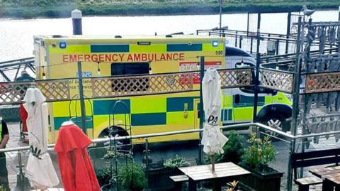 An ambulance waiting on the Quayside alongside the River Great Ouse. There are outside benches and parasols in the foreground.
