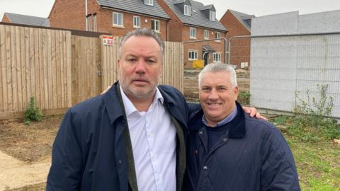 Kevin Shoemake and Dean Thomas standing side by side with their arms around each other's shoulders. Shoemake is wearing a navy raincoat which is unbuttoned and showing a white buttoned shirt underneath. Thomas is in a blue coat and smiling at the camera.