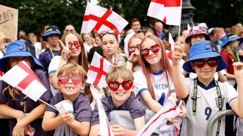 Six children standing at the front of the crowd holding England flags and wearing sunglasses with the names of Chloe Kelly and Grace Clinton written on them with pen marker. 