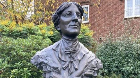 A bronze bust of a woman, she wears a high necked blouse with a dragonfly pinning it at her neck. Her hair is tied back. Her eyes gaze out to the distance. Behind the bust are bushes and a red brick building.