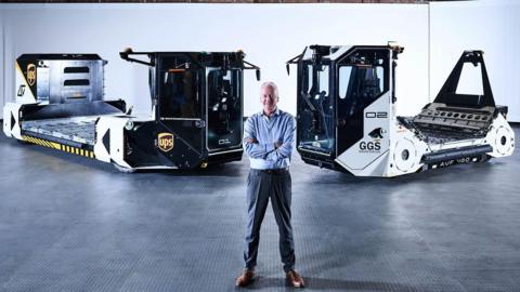 A man standing an a dark warehouse that has dim lighting. He is standing with arms crossed in front of two self-driving vehicles that are used to move airport baggage. He is wearing suit trousers and a blue shirt and has short grey hair. He is smiling. 