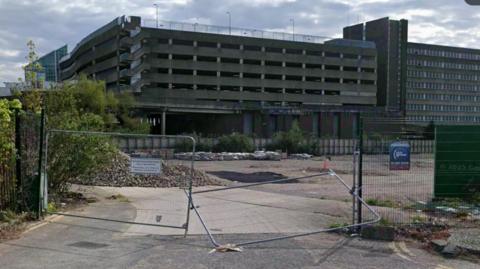 The Greyfriars site in Northampton, with metal gates across the entrance. A concrete surface is visible behind the gates, and a multi-storey car parking in brick and concrete is beyond. The sky is cloudy and grey.
