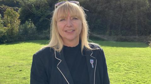 A woman with shoulder length blond hair wearing a black top and black jacket standing on a lawn with trees and bushes in the background.