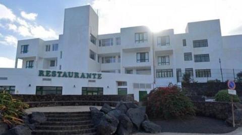 The outside of the Galeon Playa apartments with a sign for a restaurant seen in the foreground. It is a white complex, with wide steps leading up the building. 