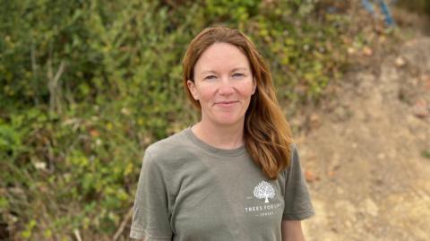 Alex Morel smiling at the camera. She has brown hair and is wearing a green T-shirt. She's stood in front of bushes.
