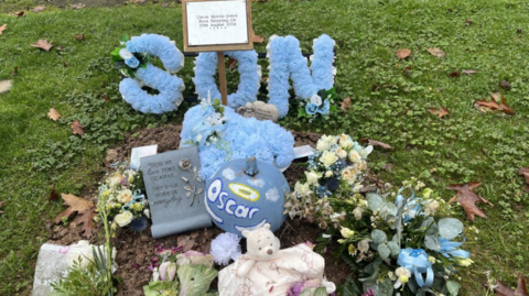 A small grave, is surrounded by grass.  There is a blue wreath that says son in blue flowers and a blue pumpkin that says Oscar with a yellow halo above the name.