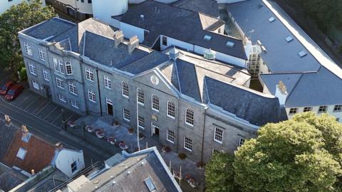 Aerial view of a large stone civic building with a slate roof, surrounded by smaller buildings and trees, with a small car park and flower planters lining the street outside.
