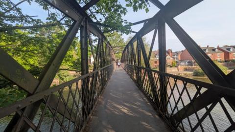 A long bridge walkway is covering a river. It is surrounded by blue sky, houses and green trees. In the long distance, we can see a person standing in the middle of the bridge.