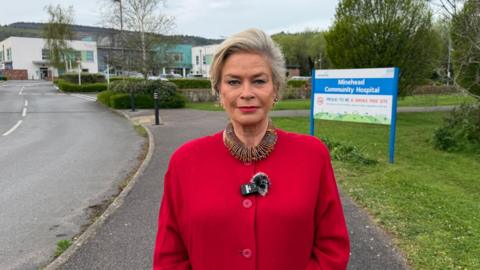 A woman stood in the middle of the frame smiling at the camera, wearing a red coat and extravagant necklace. Behind her the community hospital, and a sign directing people inside.