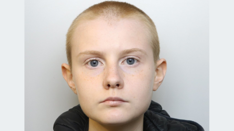 A police mugshot of Alina Burns, wearing a black hooded jacket and a grey T-shirt. She has blue eyes and her blonde hair is closely shaven. She is standing against a light grey background, looking ahead with a blank expression. 