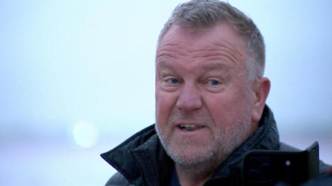 Barry Elliott has short greying hair, combed forwards from the temples, with a slight peak at the forehead and a grizzled white grey beard and slight moustache. He is wearing a black coat with the collar up. He is mid sentence, with his mouth open and forehead wrinkled and looks slightly annoyed. 