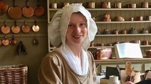 A young woman in a regency costume in a kitchen. She is wearing a beige long sleeved top with a white top underneath, a white apron and a large white hat. 