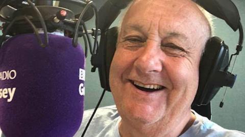 John Randall sitting behind the microphone in the BBC Guernsey studios with a big smile on his face.