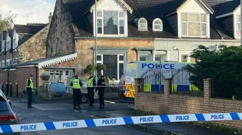 Drumoyne Road is cordoned off by police. In the foreground of the image is blue and white police tape. Four police officers can be seen standing outside a property in the background, alongside a police van.