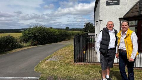 Terry and Susan Havenhand standing in front of their home which is called Railway Crossing Cottage. Terry has very short white hair and wears a white shirt, black body warmer and shorts. Susan's grey hair is tied back. She wears dark jeans, a white check shirt and yellow body warmer. They are pictured to the righ hand side of the shot with a large field, bushes and sweeping countryside on the other side.