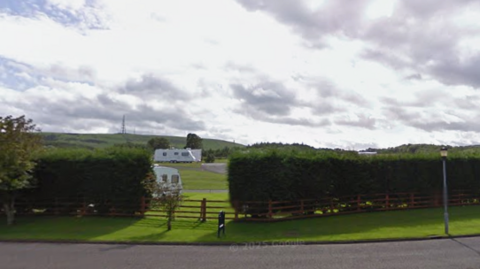 A green field with hedges at the boundary and white caravans in the distance.