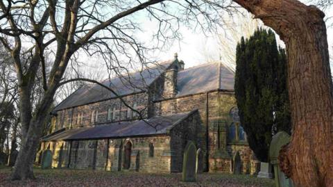 St John's church in Killingworth. The stone building sits in the grounds of an old cemetery with trees and fallen leaves on the ground.