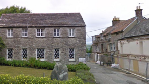 Two-storey stone buildings flank a narrow road. A traffic sign on one building says "School. Dead slow".