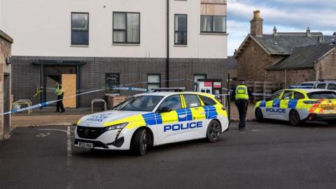 A white car with yellow and blue markings and the word police infront of a white building. Two police officers in black trousers, yellow tops and hats can be seen close to an area cordoned off with blue and white police tape. 