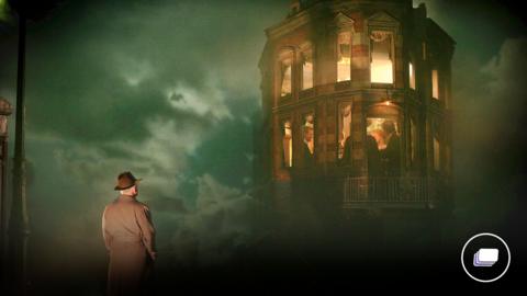 A picture from An Inspector Calls. A man is in a hat and trench coat is looking up at a tall turret with people inside. 