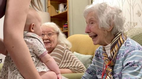 An elderly lady looks delighted to greet a six-month-old baby, who's been held by her mother. 