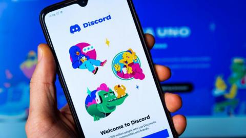 A close up shot of a person holding up a mobile phone with the login page of the Discord app shown on the screen.