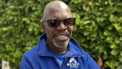 Samuel Henry wearing a blue jacket and a blue t-shirt. He is bald and wearing sun glasses. He is stood in front of a green bush.