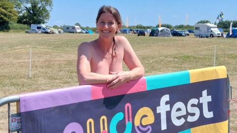 Sue poses behind a large sign for Nudefest. She is topless, and has her brown hair tied in a plait. Behind her is a green field with caravans and tents on it. 