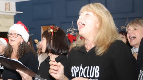 A choir is singing from song sheets in a room with a blue wall.