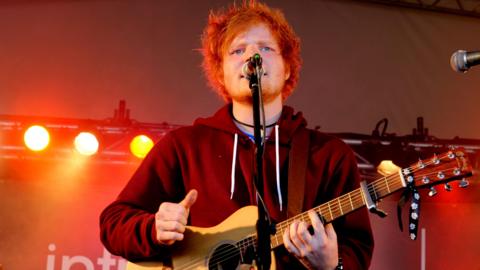 Ed Sheeran performs on the BBC Introducing Stage at Reading Festival, 2011. He has bright ginger hair and is wearing a maroon zip-up hoodie. He is singing into a microphone on a stand whilst playing an acoustic guitar.